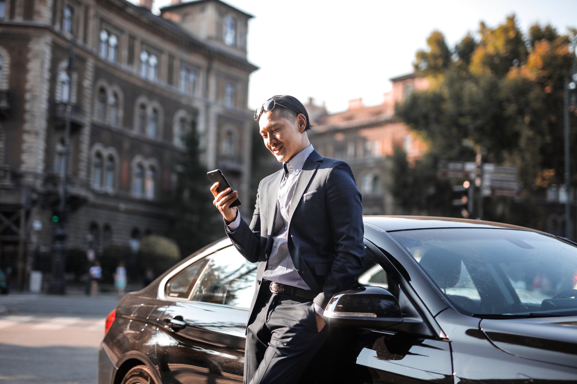 selective focus photo of man in black suit using his phone while leaning beside black car